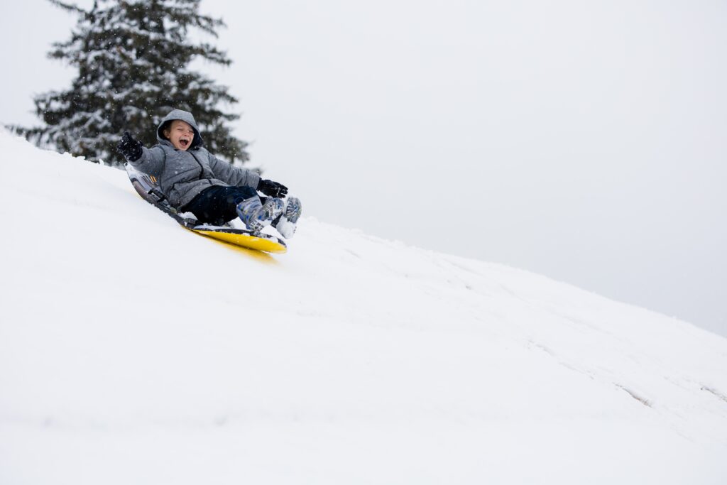 man in black jacket riding yellow snow sled on the snow-covered ground during daytime