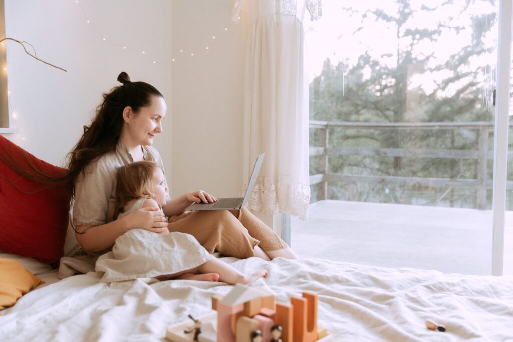 Cheerful mother cuddling daughter while sitting on bed with laptop