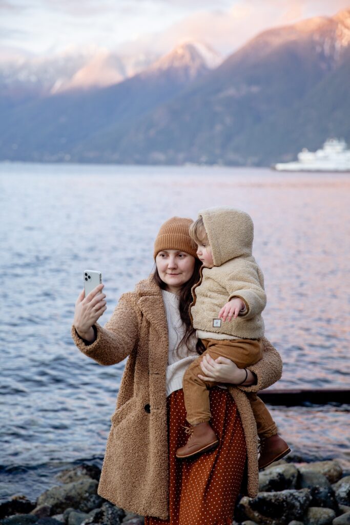 Calm woman carrying an adorable child in her hands and taking selfies on smartphones near rivers and mountains