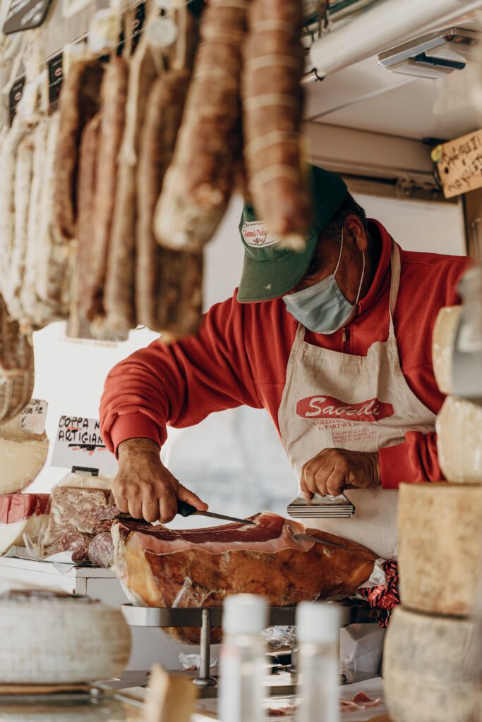 a grocer, wearing a face mask, hand cuts a local prosciutto in a shop in Sulmona, Abruzzo, Italy image