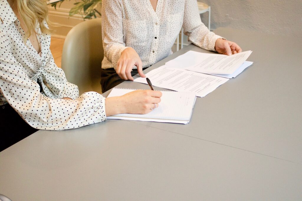 Woman signing on white paper beside woman about to touch the documents