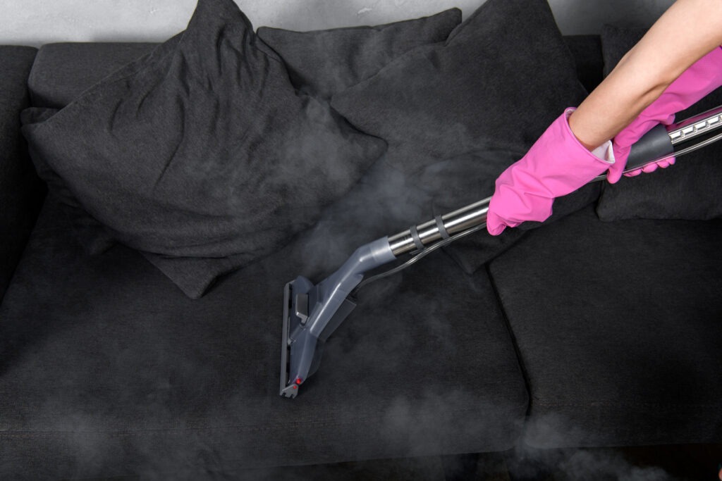 Person in rubber gloves cleaning sofa with vacuum cleaner and hot steam