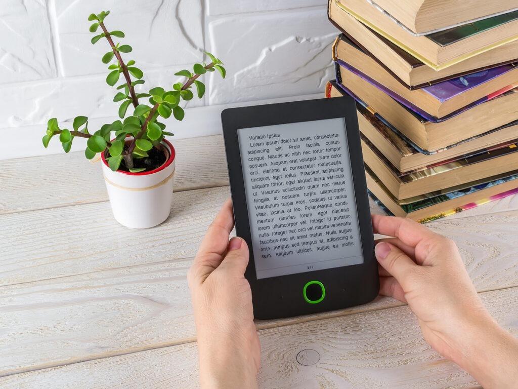 Woman hands hold e-reader near stack of paper books and small potted plant on a white wooden table