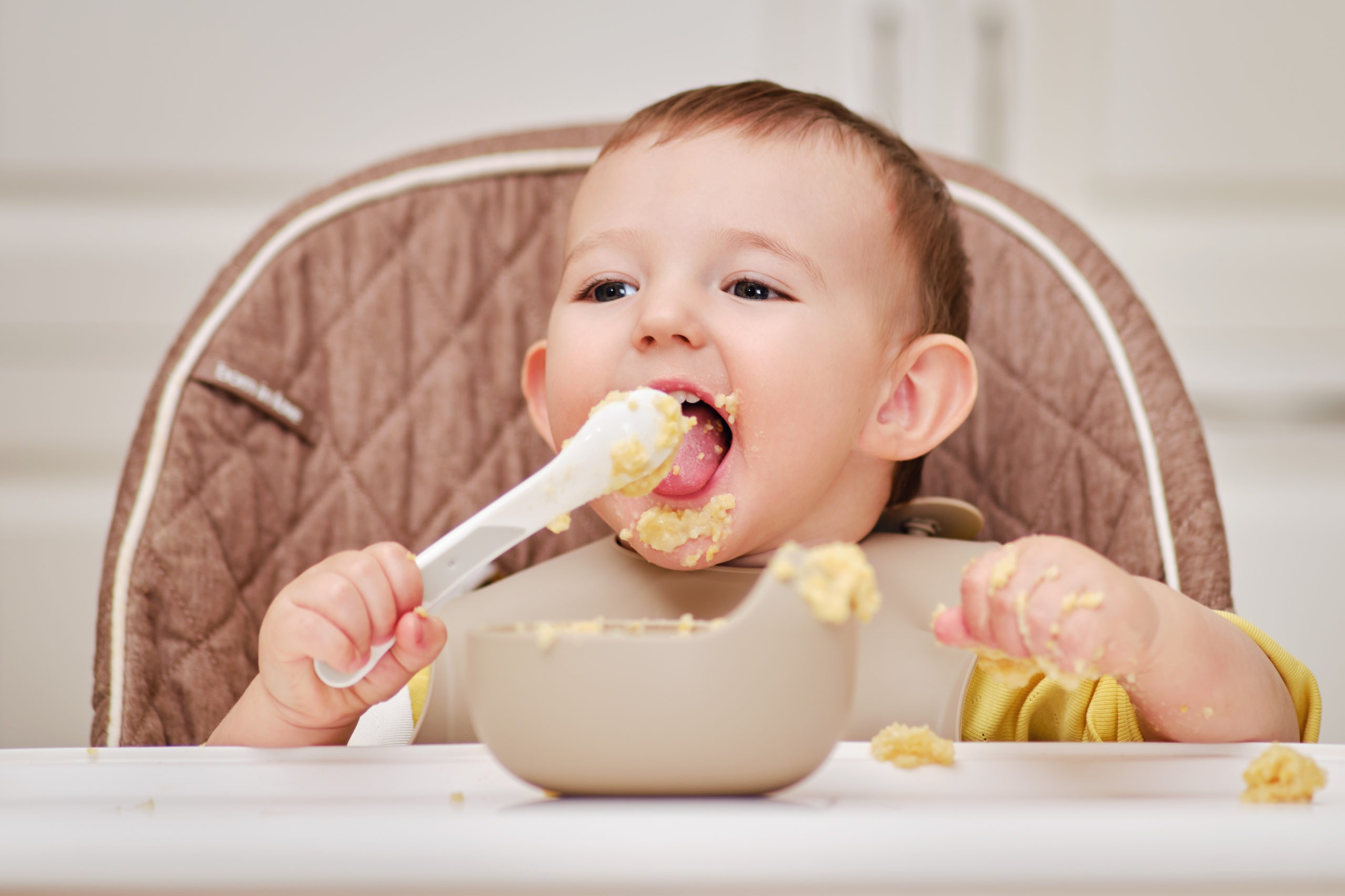 Happy toddler baby boy learns to eat porridge himself with a spoon while sitting in a child chair. Smiling child eats with his hands and a spoon from a plate, kid aged one year