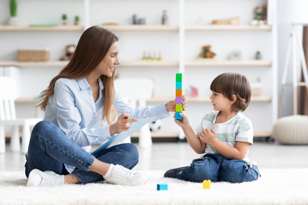 Cute little boy playing with professional female teacher, sitting together on floor at nursery and building colorful tower, empty space