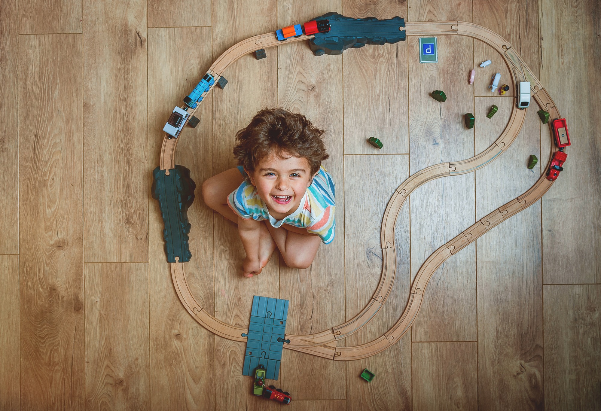 Top view of little kid boy playing wooden train set. Child sitting on floor with toys. Toys for little boy. Preschooler building rail road and blocks at home, daycare. Kindergarten educational games.