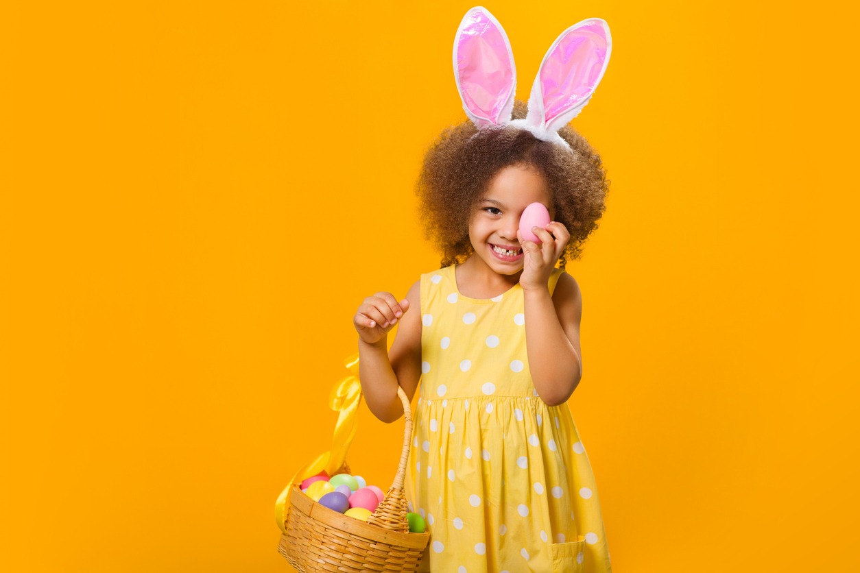 A cheerful Black girl with rabbit ears on her head with a basket of colored eggs in her hands on a yellow background