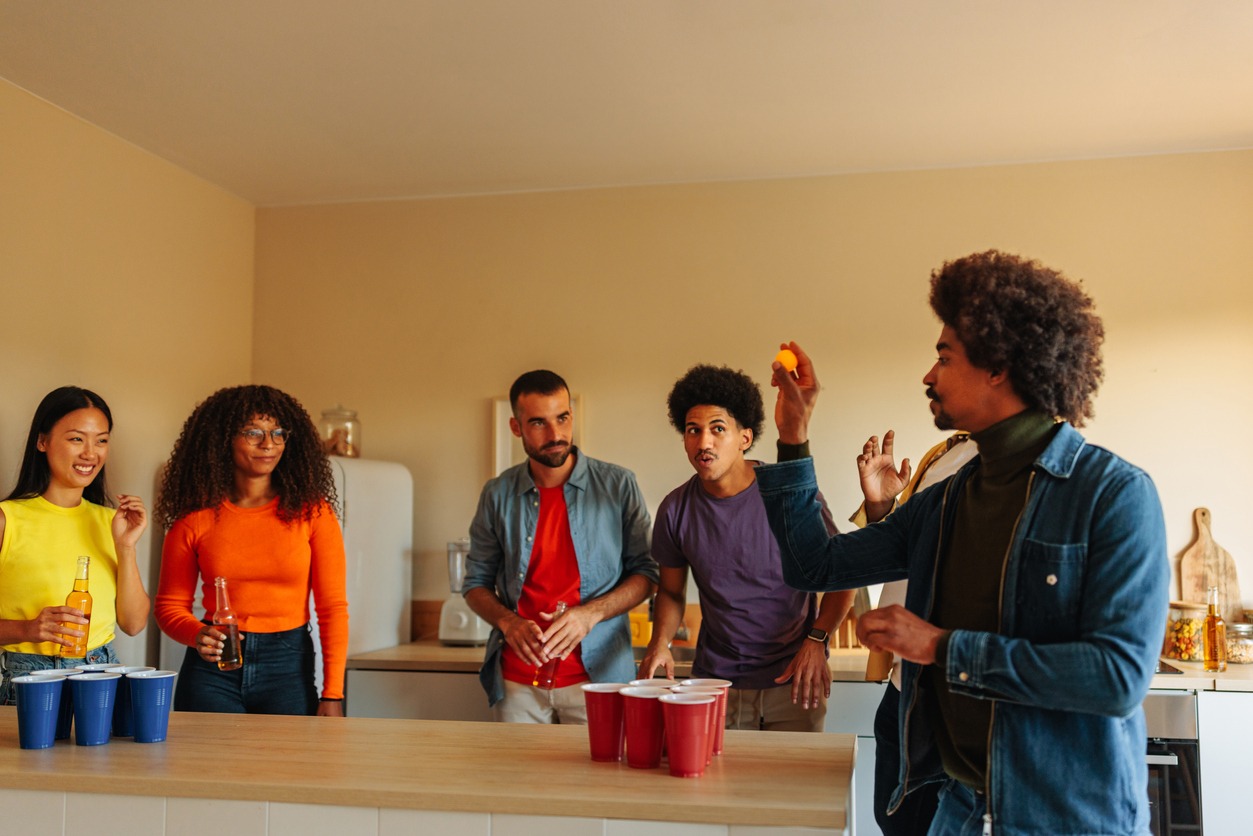 A group of young adult ethnically diverse friends are at a house party socializing over a game of beer pong in the kitchen