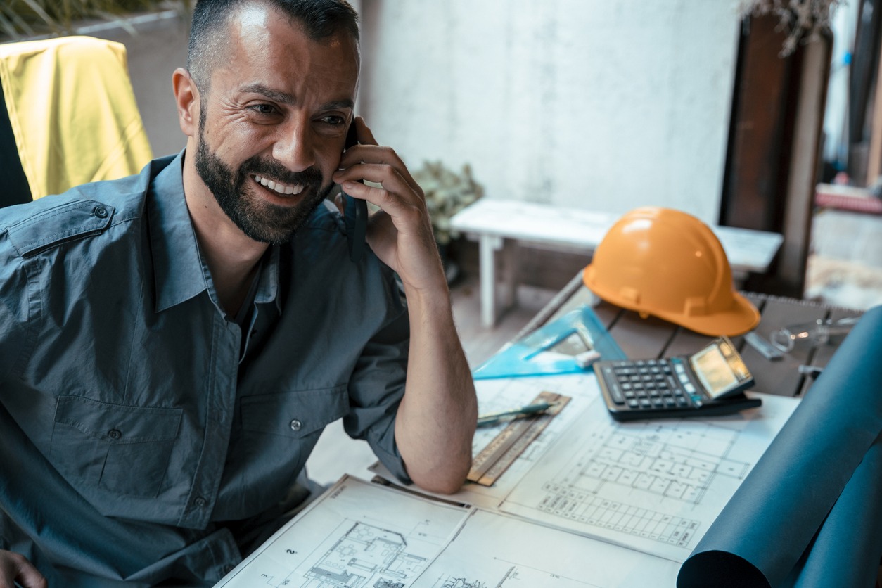a man smiling while on the phone, next to a table with blueprints and hard hat